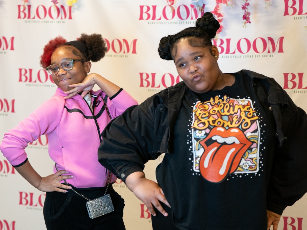 Two smiling girls pose playfully in front of a “BLOOM: Beautifully Living Out Our Me” step-and-repeat backdrop, striking confident, fun stances.